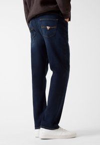 Jeans in denim blu scuro con taglio dritto, dotati di due tasche posteriori con accenti triangolari e cuciture gialle, abbinati a sneakers bianche.