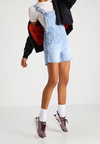 Lichtblauwe denim dungaree shorts met een rafelige zoom, gedragen over een witte geribbelde top, gecombineerd met grijze sneakers en witte sokken. Zwarte jas met rode voering.