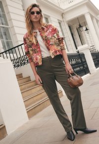 Veste bomber à motif floral en rouges et verts portée sur un haut blanc, associée à un pantalon vert olive et des talons noirs pointus. Accent sac à main marron.