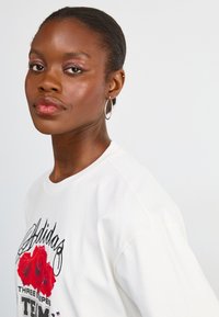 Hvid bomulds t-shirt med et sort Adidas-logo og grafisk rødt rosemotiv. Modellen bærer sølv hoops-øreringe. Blød tekstur, afslappet pasform.