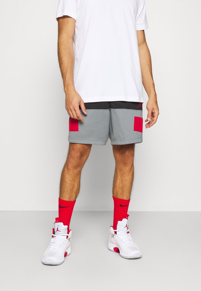 Homme portant un short gris avec des patchs rouges, un T-shirt blanc, des chaussettes rouges Nike et des baskets blanches avec des accents rouges, debout sur un sol gris.