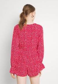 Combinaison rouge à pois blancs avec manches longues, poignets à volants et taille cintrée. Confectionnée dans un tissu léger, elle comporte une fermeture à boutons au dos.