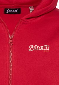 Sudadera con cremallera roja y capucha, con un logotipo blanco en contraste de "Schott NYC", textura suave y cremallera de metal. Etiqueta de tamaño visible.