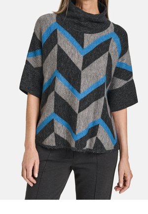 Nainen, joka pukeutunut harmaaseen, mustaan ja siniseen vinokuviolliseen lyhythihaiseen poolokaulukselliseen neulepaitaan ja mustiin housuihin.