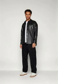 Άντρας που φοράει ένα μαύρο ψεύτικο δερμάτινο πουκάμισο με κουμπιά πάνω από ένα λευκό μπλουζάκι, μαύρο μοτίβο παντελόνι και λευκά αθλητικά παπούτσια, χωρίς φόντο.