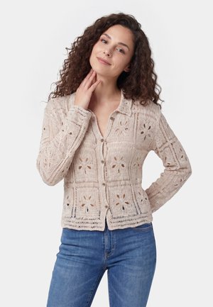 Kvinde med krøllet brunt hår iført en beige hæklede cardigan med knapper og blå jeans, der rører ved sin nakke og smiler blidt.