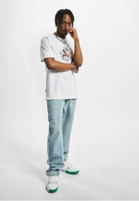 Weißes Grafik-T-Shirt mit buntem, kreisförmigem Design; kombiniert mit hellblauen Jeans und weißen Sneakern mit grünen Akzenten.