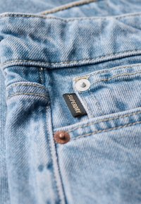 Tela de denim azul con un lavado ligero. Presenta una etiqueta oscura con "Superdry", un botón a presión plateado y detalles de costura visibles.