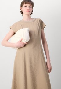Beige dress with a textured finish, short sleeves, and a round neckline. Model holds a cream-colored, folded garment against the chest.