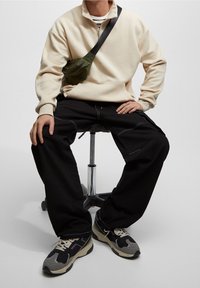 Beige sweatshirts med lynlås, sorte løstsiddende bukser, grå sneakers med lilla detaljer og olivengrøn skuldertaske med lynlåsdækning.