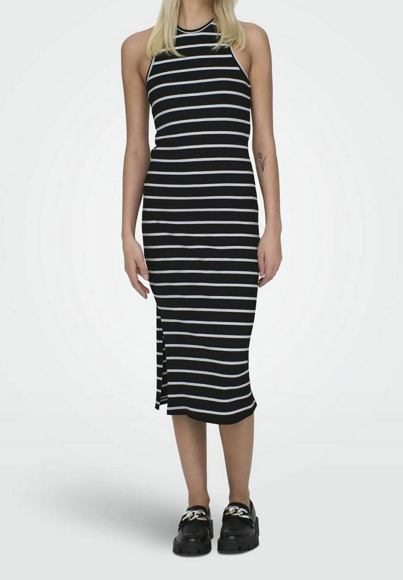 Femme portant une robe midi noire sans manches avec des rayures horizontales blanches et des mocassins noirs épais avec un détail chaîne argent.
