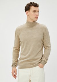 Beige Rollkragenpullover aus weichem Material, enganliegendes Design, gerippte Bündchen und Kragen. Das Modell posiert mit einer Hand in der Tasche vor neutralem Hintergrund.