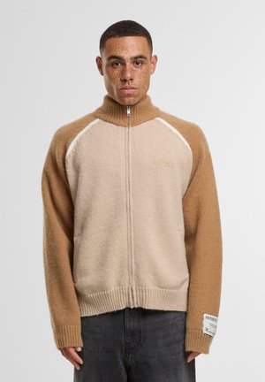 Gilet zippé beige et tan avec col montant, poignets côtelés et finition texturée. Présente des manches raglan contrastées et un détail de tag.