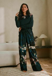 Dunkelgrünes Langarmoberteil mit floralem Design am Ausschnitt, kombiniert mit weiten schwarzen Hosen mit großen Blumenmustern. Weiche Textur.