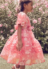 Roza cvetna obleka s kratkimi napihnjenimi rokavi, slojenimi volani in dekorativnim cvetom na pasu. Prosojen material z nežnim sijajem.