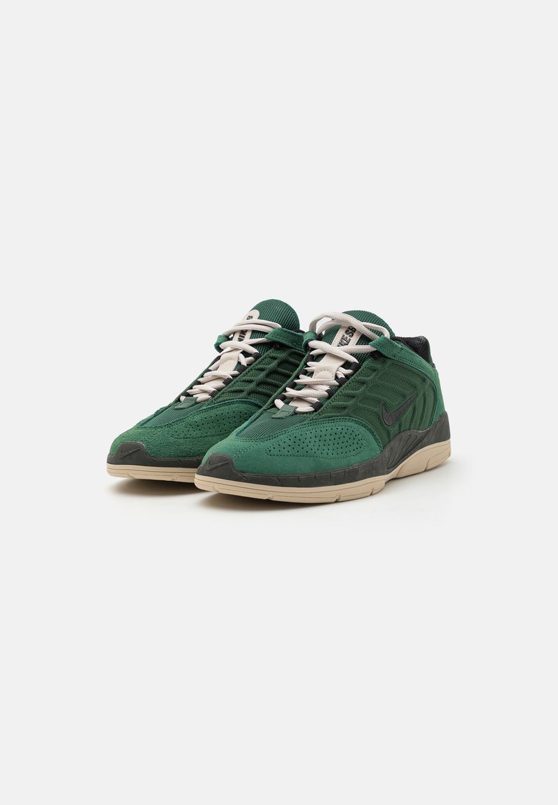 Zapatillas de gamuza verde con paneles de malla, cordones crema y un acento negro. Presentan una suela texturizada y un diseño de puntera redondeada.