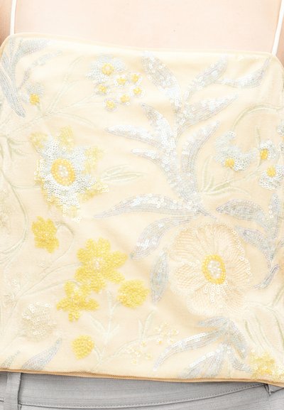 Κίτρινη τοπ από δίχτυ με κεντήματα λουλουδιών σε μπλε και κίτρινες αποχρώσεις, στολισμένη με παγιέτες, δημιουργώντας ένα υφασμένο, περίπλοκο μοτίβο.