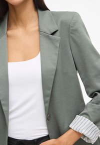Lichtgroene blazer met een gestructureerde kraag, knoopdetail en opgerolde mouwen met een wit gestreept patroon. Draagt over een witte top.