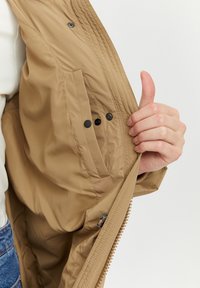 Tan lightweight jacket with a zip closure, featuring a hidden pocket and snap buttons. The fabric is smooth with a subtle sheen.
