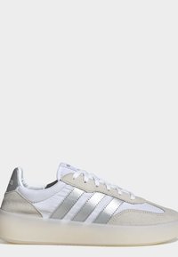 Basket blanche profil bas avec des empiècements en daim gris clair, trois bandes métalliques argentées, lacets blancs et semelle beige translucide.