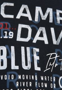 Črna bombažna majica s poudarjenim belim in rdečim besedilom, ki vsebuje različne fraze, vključno z "CAMP DAVID" in "IZOGIBAJTE SE GIBLJIVI VODI."