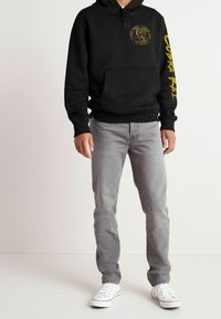 Sudadera negra con capucha, con texto amarillo y gráfico de una cobra; combinada con jeans ligeros grises y zapatillas blancas. Tela suave, corte casual.