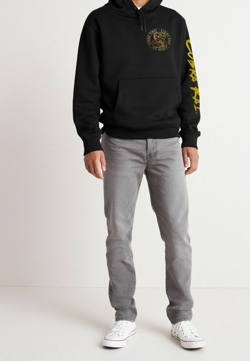 Sudadera negra con capucha, con texto amarillo y gráfico de una cobra; combinada con jeans ligeros grises y zapatillas blancas. Tela suave, corte casual.