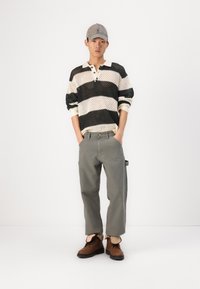 Csíkos kötött pulóver fekete és krém színben, szürke nadrág és barna cipő. A sapka szürke, logóval ellátva. Az outfit laza, kényelmes fazonnal rendelkezik.