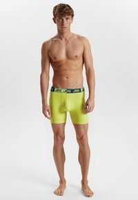 Neon gul boxer shorts med en blå og grøn mærkebånd, der har en tætsiddende pasform og glat tekstur. Modellen står barfodet og uden skjorte.
