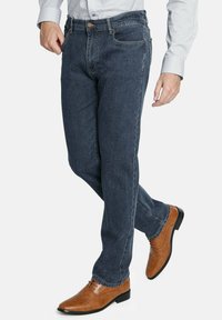 Jeans de denim azul oscuro con un diseño de pierna recta, que cuentan con un cierre de botón de cobre y bolsillos delanteros, combinados con zapatos de vestir de cuero marrón.