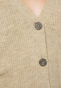 Beige gebreide cardigan met een V-hals, voorzien van twee donkere knopen en een getextureerd oppervlak. Zichtbare stikpatronen versterken het ontwerp.