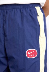 Marineblaue Sporthosen mit elastischem Bund, hellbeige Seitenstreifen und einem roten Nike AIR-Logo auf dem Oberschenkel. Glattes, leichtes Material.