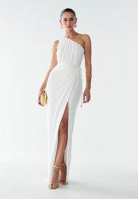 Vit, axelbandslös klänning i mjukt tyg; har en omlottsdesign med hög slits på låret och ett knyte i midjan. Guldaccesoarer som detaljer.