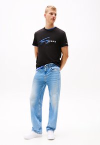 Black t-shirt with blue accents and logo, paired with light blue distressed jeans. Model wears white sneakers. Simple, casual attire.
