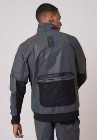 Giacca impermeabile iridescente con colletto alto, dettagli neri e tasca con zip sulla schiena. Realizzata in materiale leggero.