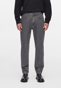 Vaqueros de mezclilla gris con corte recto, diseño de cinco bolsillos y cierre de botón, usados con una camisa negra de manga larga y botas negras.