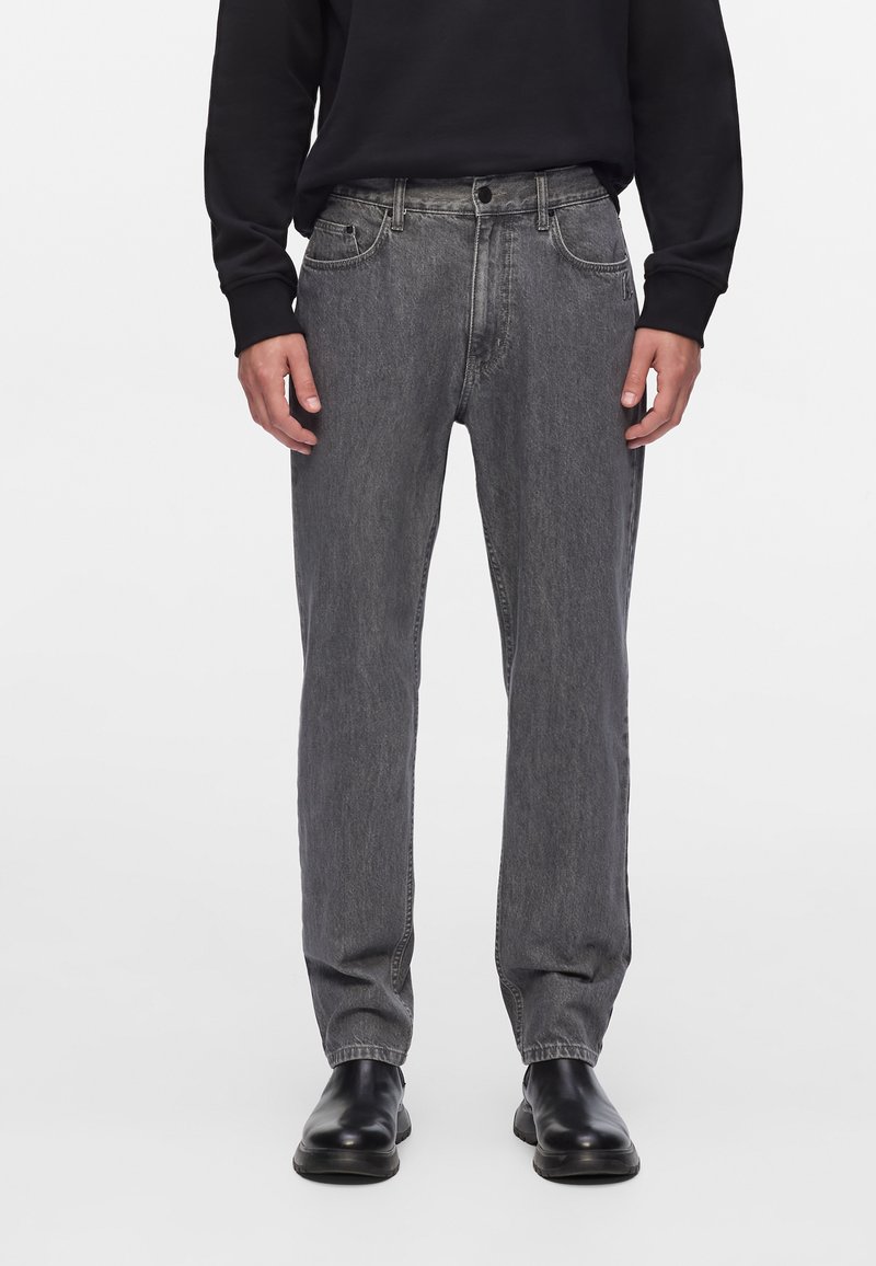 Vaqueros de mezclilla gris con corte recto, diseño de cinco bolsillos y cierre de botón, usados con una camisa negra de manga larga y botas negras.