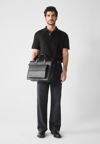 Maletín de piel negra con asas superiores y correa ajustable, que cuenta con un bolsillo frontal y un diseño estructurado. El modelo lleva pantalones oscuros y una polo.