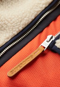 Jacka med ett mysigt beige fleecetyg på insidan, livlig orange meshyta, mörkblå detaljer och en läderbricka med "SUPERDRY"-märke.