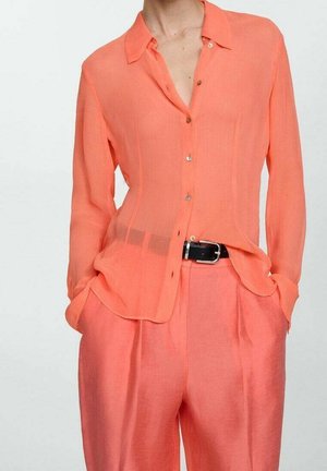 La personne porte une chemise corail transparente boutonnée, rentrée dans un pantalon taille haute assorti, les mains dans les poches et une ceinture noire à la taille.