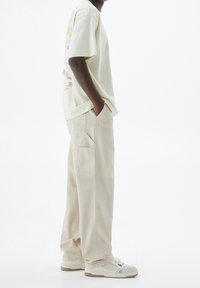 T-shirt oversized jaune clair avec design graphique, associé à un pantalon cargo beige doté de poches, et des baskets claires avec des accents blancs et gris.