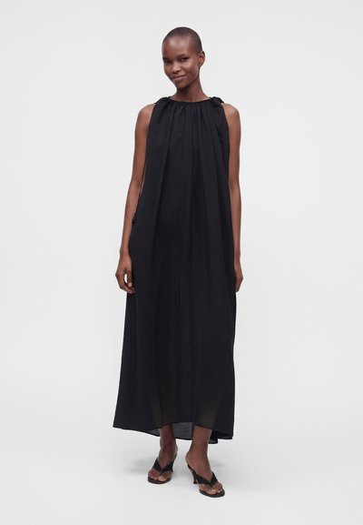 Femme adulte debout portant une longue robe noire sans manches avec un col froncé et des sandales noires à talons sur un fond clair uni.