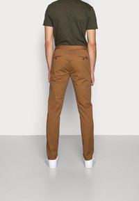 Pantalon en coton marron au design droit, avec deux poches arrière et un détail en bouton. Porté avec un t-shirt gris foncé et des chaussures blanches.