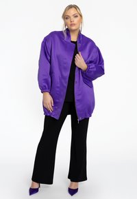 Veste bomber en satin violet avec fermeture éclair, poignets élastiques et poches latérales ; portée sur un top noir et associée à un pantalon noir.