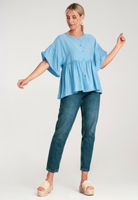 Jasnoniebieska bluzka z krótkim rękawem, zapinana na guziki, z falbaniastym dołem i luźnym krojem, zestawiona z ciemnoniebieskimi dżinsami i beżowymi sandałami na platformie.