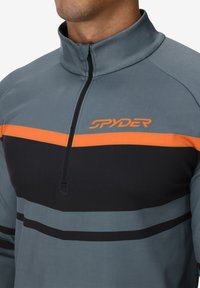 Pullover grigio con colletto alto, caratterizzato da una trama orizzontale arancione e nera. Il materiale appare liscio ed elastico. Logo sul petto.