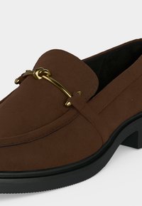 Mocassin en daim marron avec détail en métal doré et semelle en caoutchouc noire, doté d’un petit talon et d’un design slip-on.