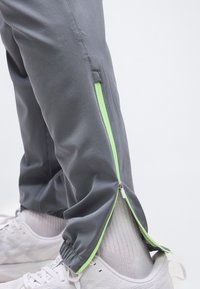 Κοντινό πλάνο γκρι παντελονιού με πράσινο φερμουάρ μισάνοιχτο πάνω από λευκά αθλητικά παπούτσια και κάλτσες.