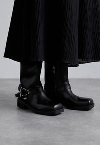 Bottines en cuir noir avec un bout carré, des détails de sangle, des accents métalliques et un petit talon épais, associées à une jupe plissée noire.