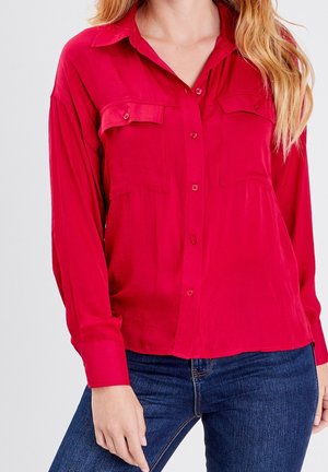 Femme portant une chemise rouge à manches longues boutonnée avec des poches poitrine et un jean bleu, debout devant un fond clair uni.